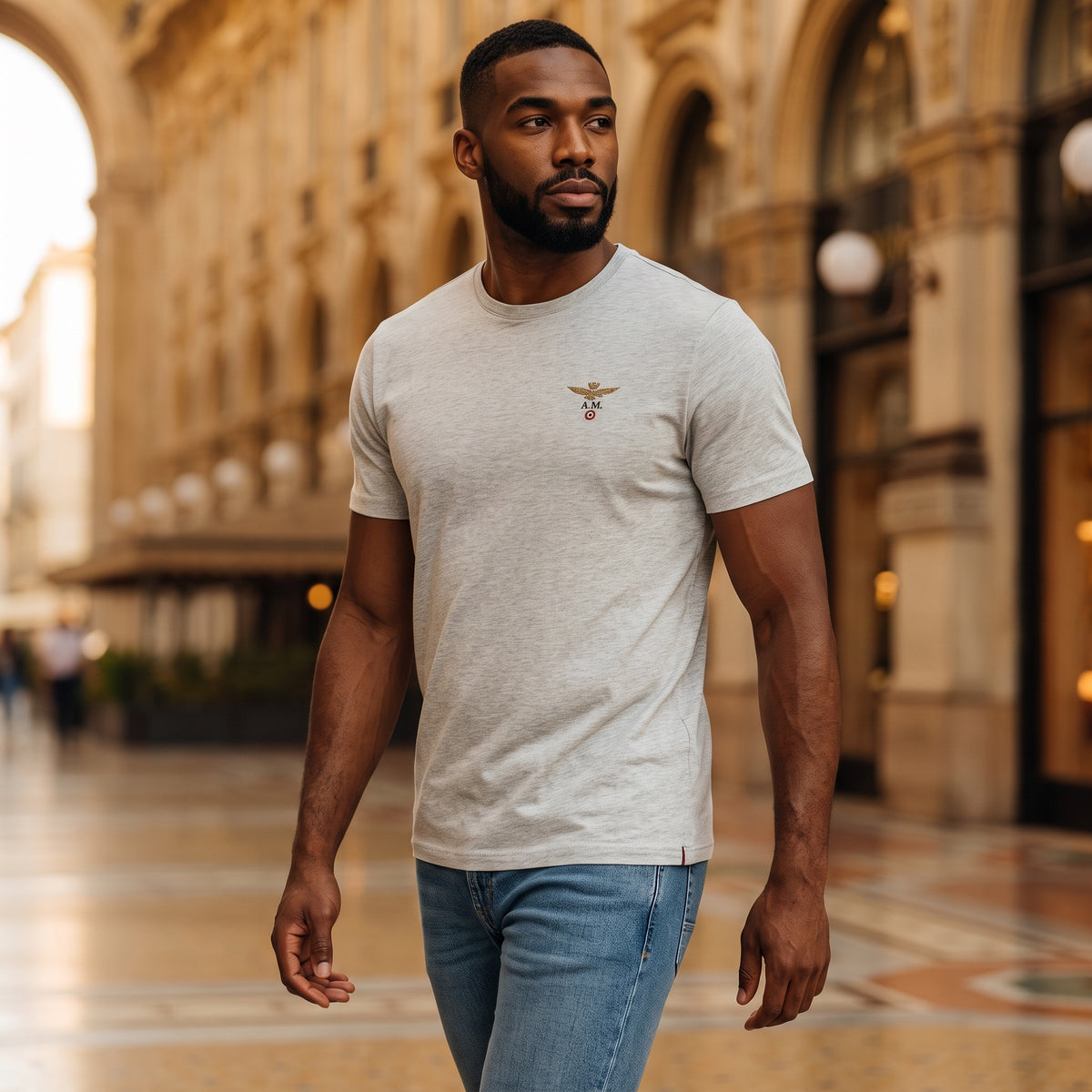 Hombre con camiseta gris de cuello redondo Aeronautica Militare y logo de alas en el pecho en entorno urbano