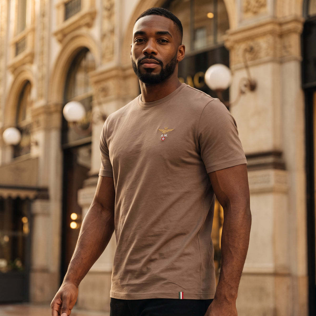 Hombre vistiendo camiseta marrón de cuello redondo Aeronautica Militare con logo de alas en el pecho en entorno urbano