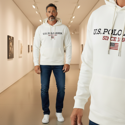 Hombre con sudadera blanca con capucha U.S. Polo Assn. y logo bordado, combinada con vaqueros oscuros en una galería